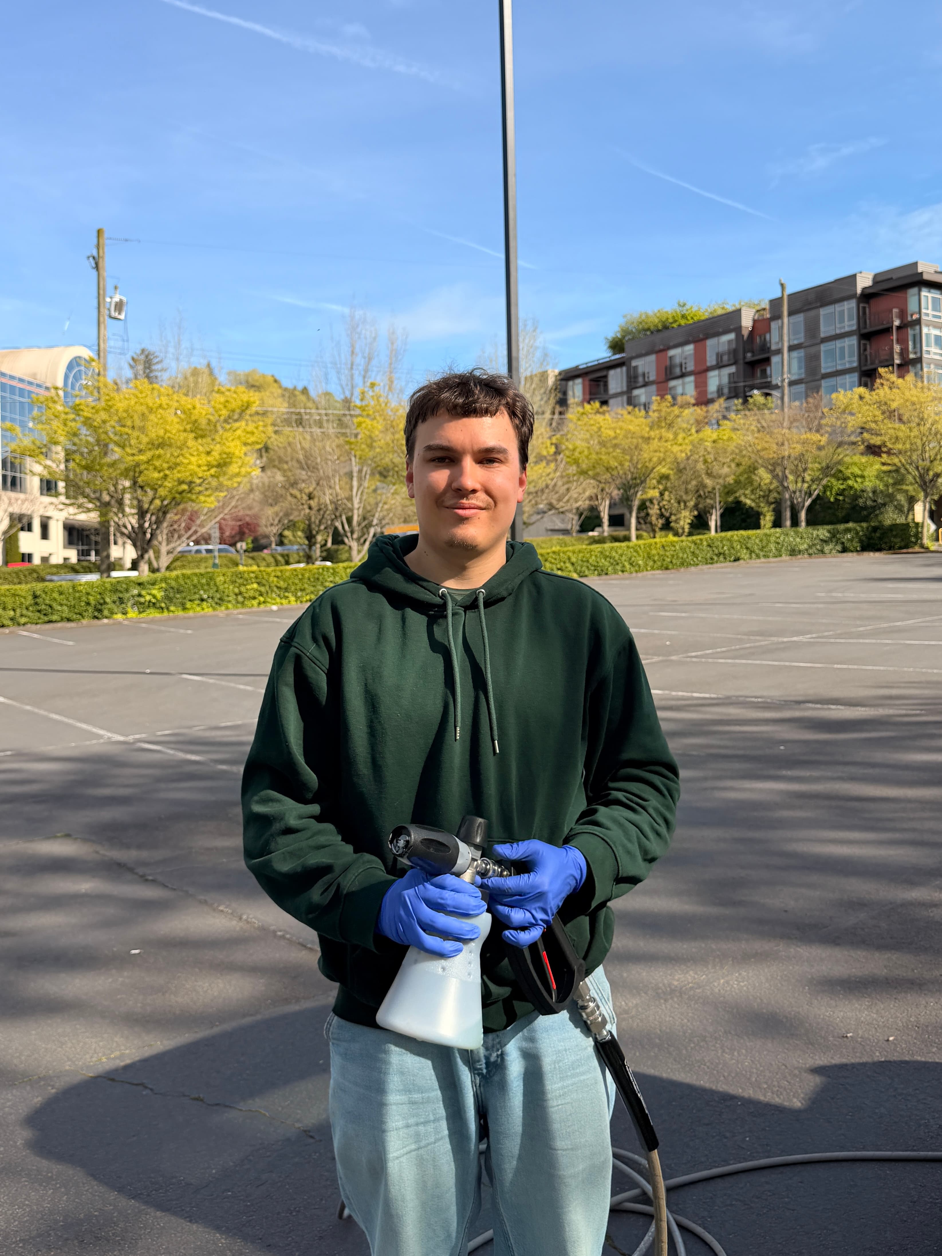 Man in a green hoodie holding a foam cannon attachment in a sunny parking lot.